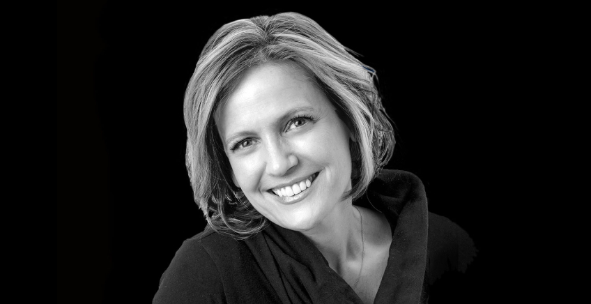 Portrait of a smiling woman with short blonde hair wearing a black top against a dark background.