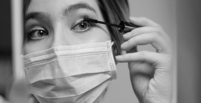 Close-up of a person wearing a surgical mask while applying mascara to their upper eyelashes.