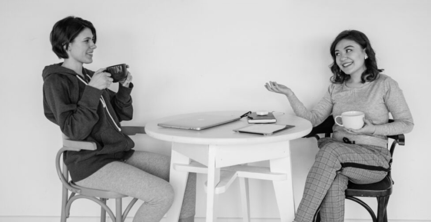 Two people sit at a round table with laptops and mugs, smiling and talking.