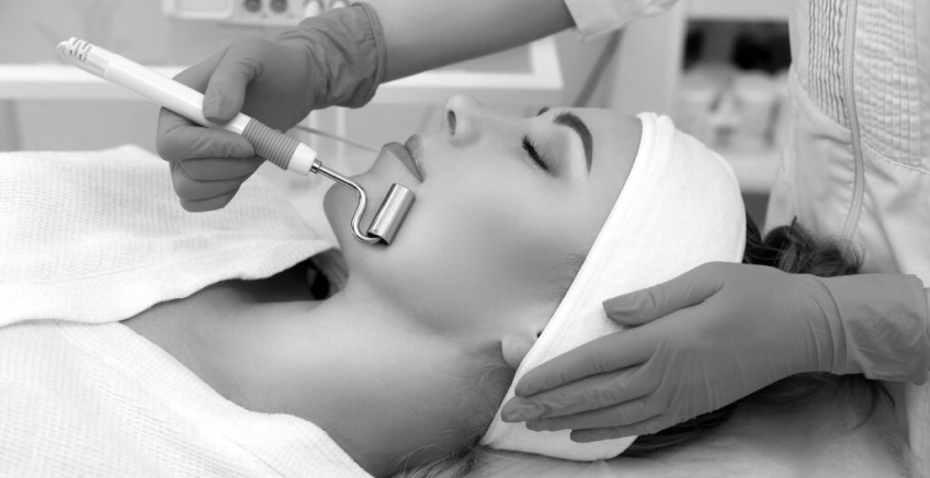 Patient lying with a headband during a facial treatment; clinician in gloves uses a metal wand near her mouth for a cosmetic procedure.