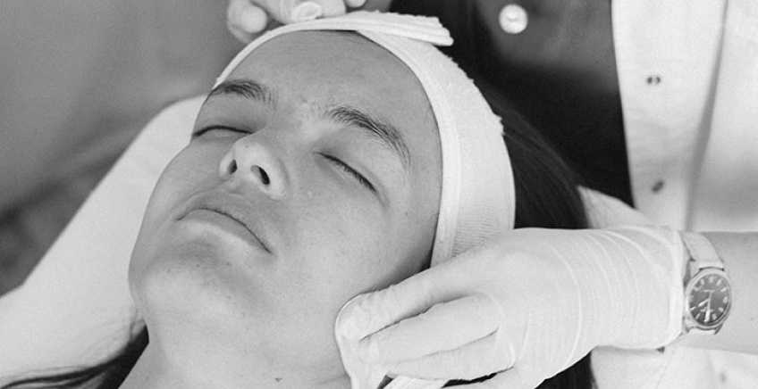 Woman lying with head wrapped, eyes closed, receiving a facial treatment from gloved hands.