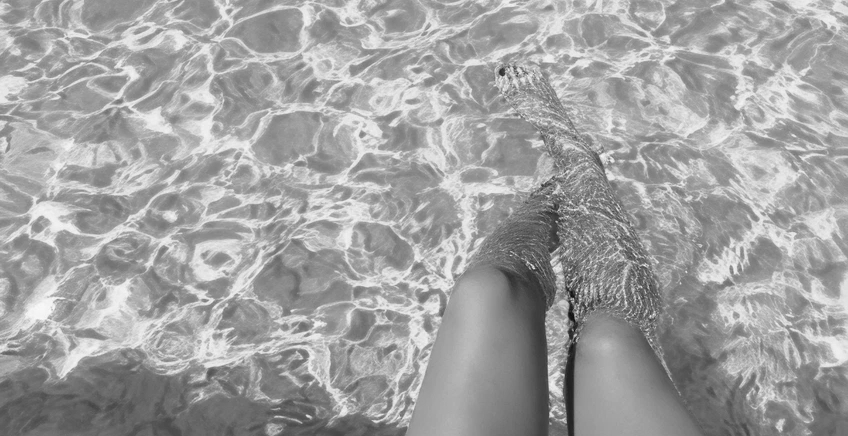 Person's legs submerged in shallow, sunlit water with rippled patterns on the surface.