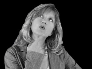 Woman in a pinstripe blazer gazes upward, finger at chin in thoughtful pose against a black background (black-and-white photo)