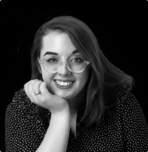 Portrait of a smiling woman with round glasses, resting her chin on her hand in a black-and-white photo, wearing a dotted blouse.