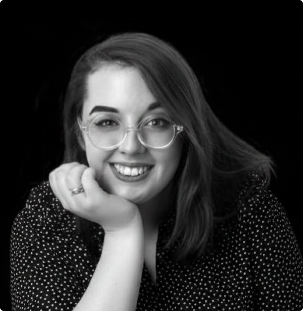 Portrait of a smiling woman with round glasses, resting her chin on her hand in a black-and-white photo, wearing a dotted blouse.