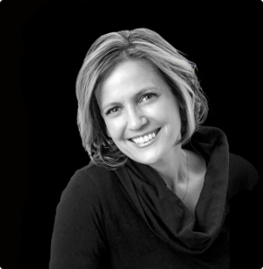 Black-and-white head-and-shoulders portrait of a smiling woman with short light hair, wearing a black top with a draped neckline, against a dark background