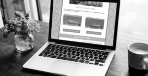 Monochrome photo of a laptop on a wooden table, with a vase of flowers on the left and a mug on the right, displaying a promotional webpage on screen.