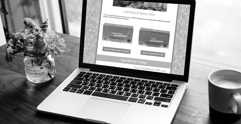 Monochrome photo of a laptop on a wooden table, with a vase of flowers on the left and a mug on the right, displaying a promotional webpage on screen.